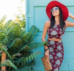 stylish beautiful woman in red hat posing on blue background, printed outfit, summer style, fashion trend, top, skirt, skinny, straw handbag, sunglasses, accessories, smiling, happy, tropical vacation